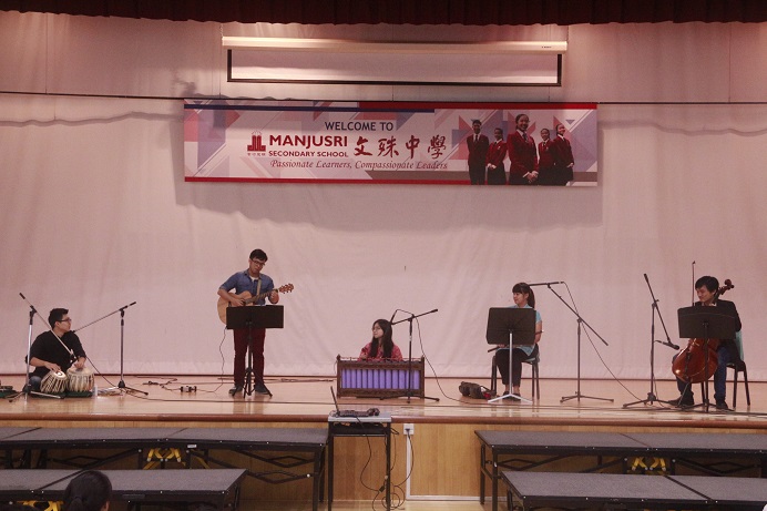 Music ensemble performing on a school stage with instruments including guitar, drums, and cello.
